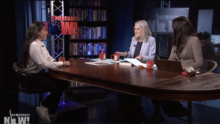 Shreya Johri on the Democracy Now! set, seated at the studio table across from the two hosts, with the show's logo and the newsroom behind them.